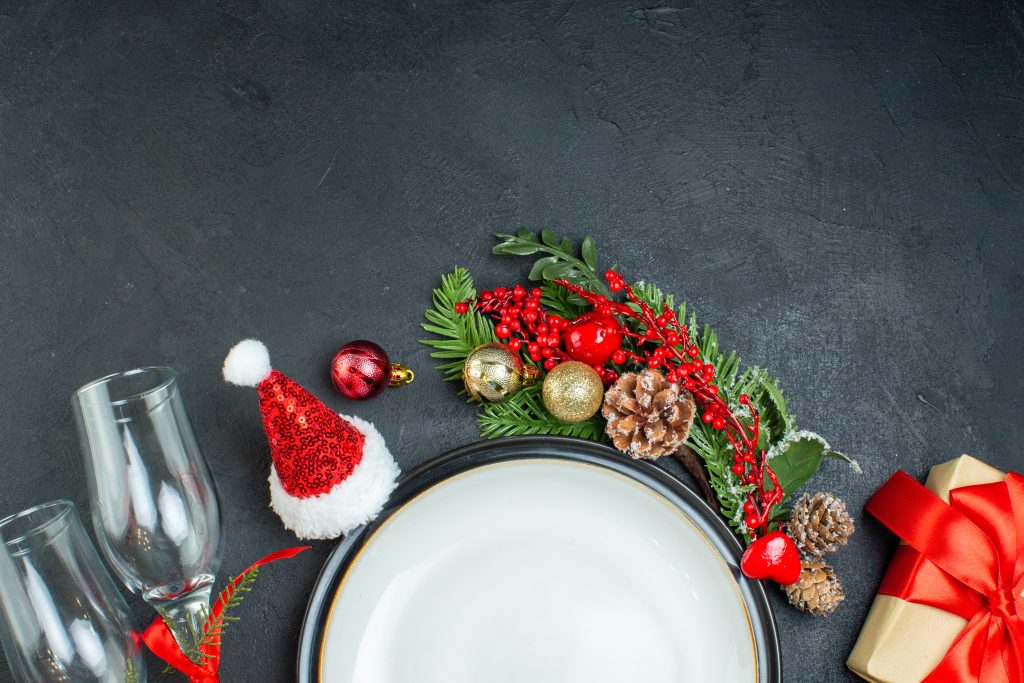 half-shot-dinner-plate-christmas-tree-fir-branches-conifer-cone-gift-box-santa-claus-hat-fallen-glass-goblets-black-background